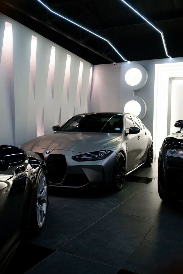 Grey BMW M3 Competition with its large kidney grille, parked between darker cars in a showroom with lit white panels