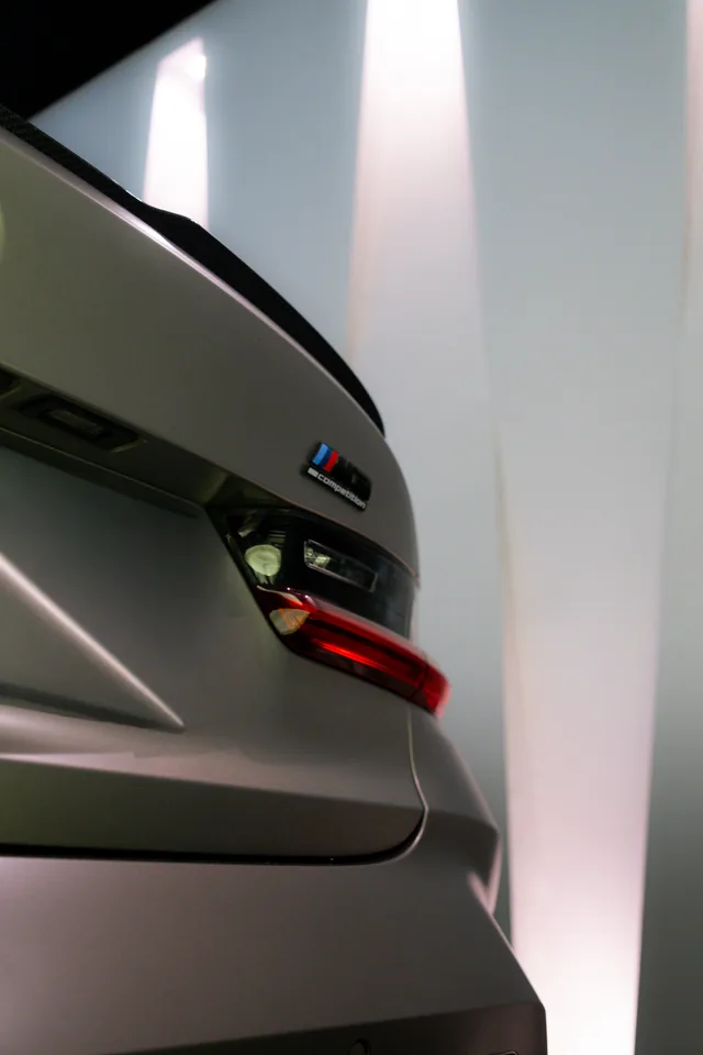Rear corner of a matte grey BMW M3 with its M badge and tail light, lit by a pale showroom wall