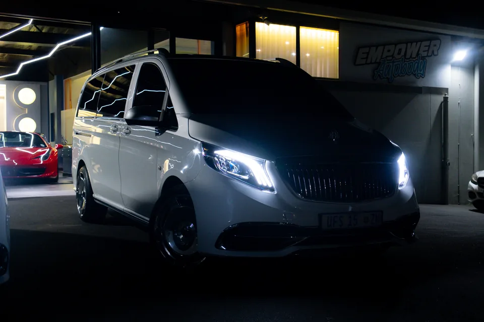 White Mercedes-Benz van with its headlights on outside a showroom at night, a red sports car visible inside