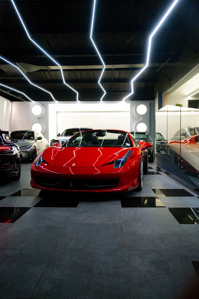 Red Ferrari 458 Spider in the centre of a showroom beneath zig-zag ceiling lights, with more cars lined up behind