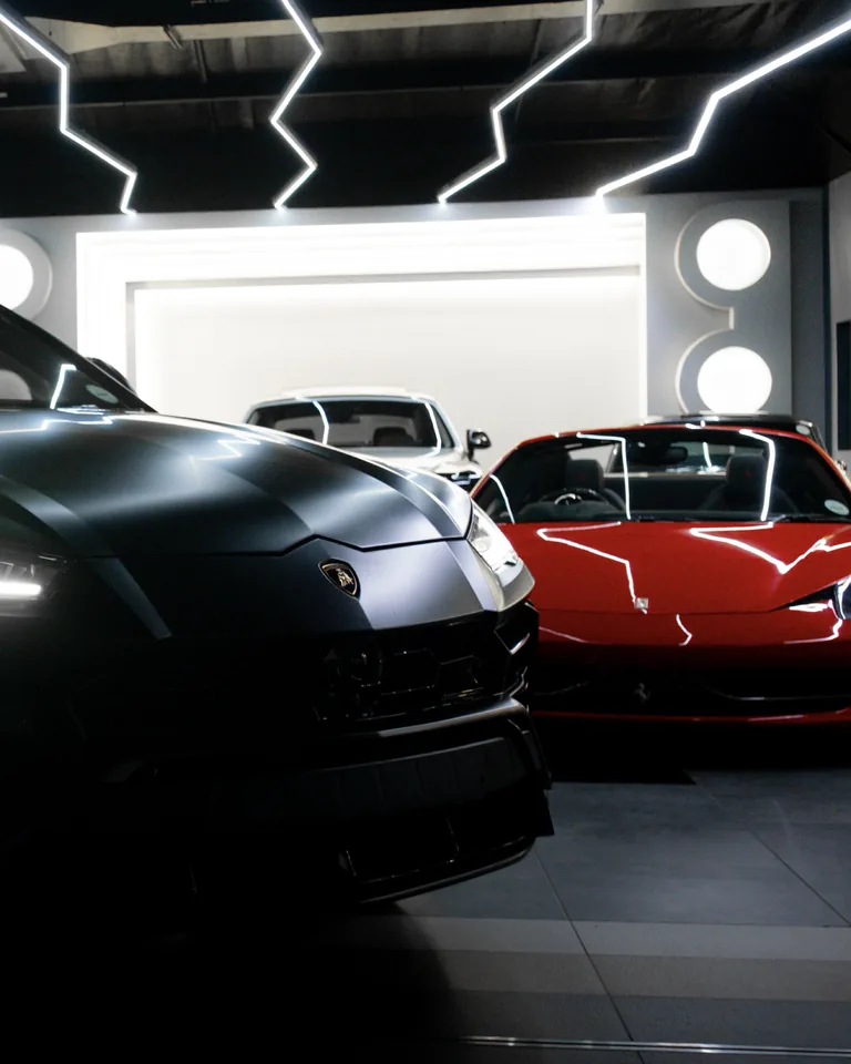 Matte grey Lamborghini Urus in the foreground with a red Ferrari 458 Spider beside it under lightning-shaped ceiling lights