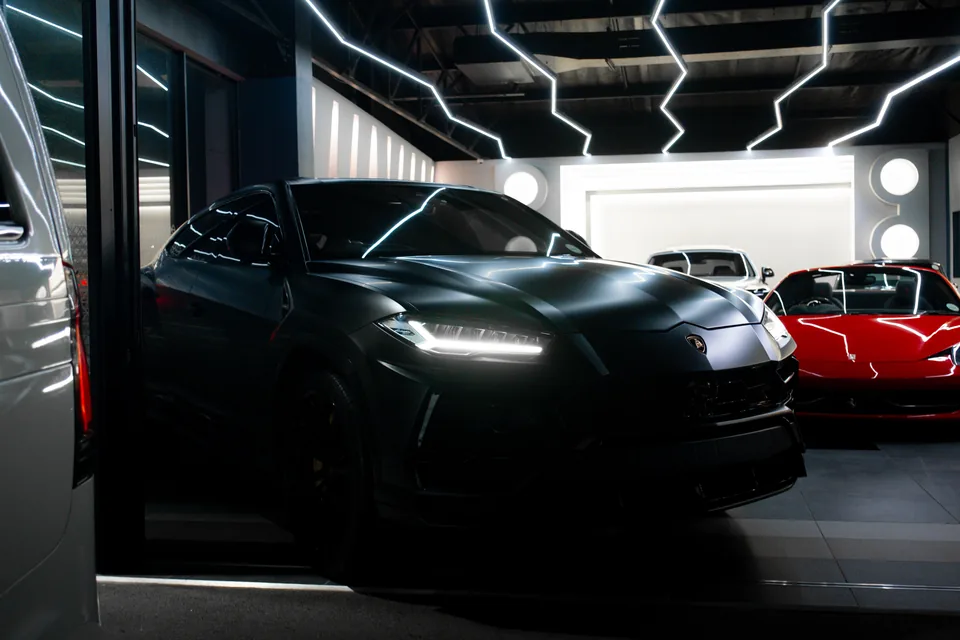 Matte grey Lamborghini Urus with its daytime running lights on, parked in a showroom under zig-zag ceiling lights, a red Ferrari beside it