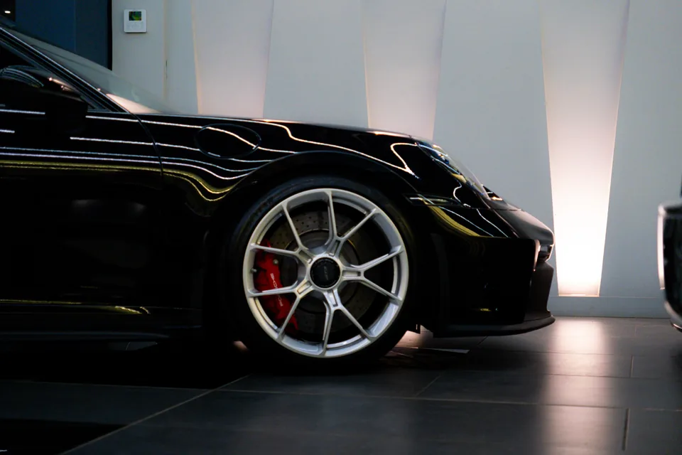 Front of a black Porsche 911 GT3 with a silver centre-lock wheel and red brake caliper against a backlit white wall
