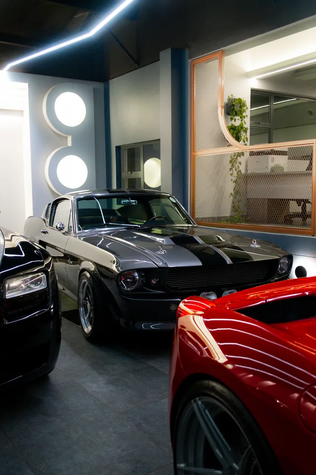Grey 1967 Shelby GT500 parked in the showroom with the rear of a red Ferrari in the foreground and a Rolls-Royce nose to the left